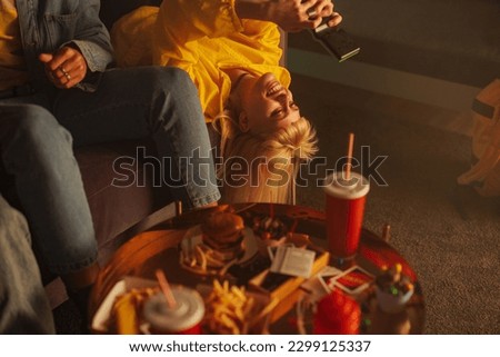 A cheerful stylish woman is lying on a sofa upside down with her friends at a retro party and playing a vintage video game.