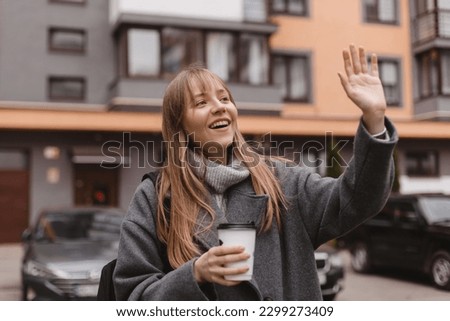 Smiling young woman waving her hand with a friendly cheerful smile to her neighbours. Girl wear grey sweater and coat, hold cup of tea, meeting friends, welcome, nice to meet you. Hi, hello gesture.  Royalty-Free Stock Photo #2299273409