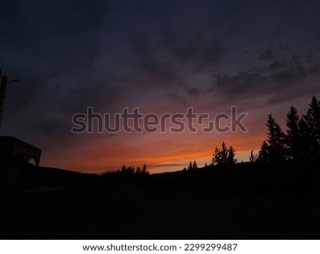 "A picture of a sunset from one of the mountainous areas in a magnificent view with the reddening of the sky that added a magical touch to the image, and darkness below with trees and the mountain."