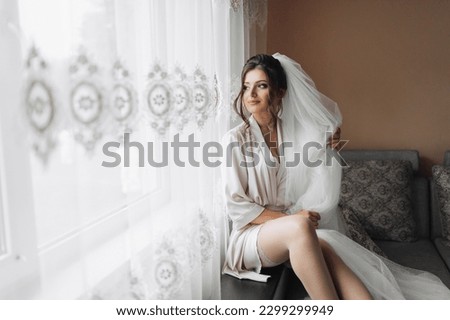 Portrait of the bride. A brunette bride is sitting on a gray sofa in a dressing gown, posing, holding her voluminous white veil. Gorgeous make-up and hair. Wedding photo. Beautiful bride