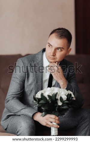 Portrait. A man in a white shirt, black tie and gray suit is sitting on a sofa, holding a bouquet of white roses. A stylish watch. Men's style. Fashion. Business