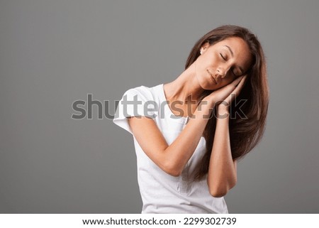 Beautiful young woman with long hair mimics sleeping gesture, hands joined, pretending to sleep on hand-formed pillow, tilting head to one side, conveying calmness and serenity Royalty-Free Stock Photo #2299302739