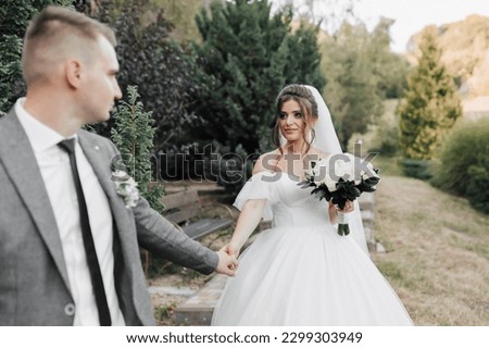 portrait of the bride and groom in nature. Stylish groom and brunette bride in a white voluminous dress, walking, holding hands and looking at each other against the background of the forest.