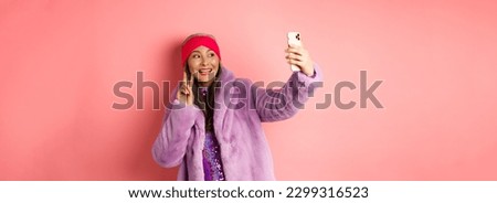 Fashion concept. Cool and cheerful asian senior woman taking selfie with peace sign, wearing trendy faux fur coat and party dress, standing over pink background.