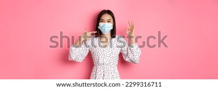 Coronavirus, social distancing and lifestyle concept. Cute asian woman pointing at face mask, showing okay sign, support using preventive measures from covid-19, pink background.