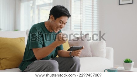 Asia people young man relax sitting easy at home sofa couch laugh smile enjoy play video game social media on mobile app, win online stream casino battle trade, leisure teen activity happy fun joy. Royalty-Free Stock Photo #2299437089