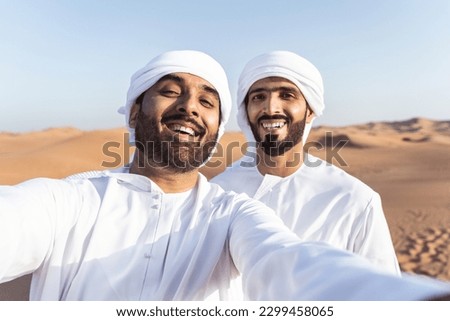 Two middle-eastern men wearing traditional emirati arab kandura bonding in the desert - Arabian muslim friends meeting at the sand dunes in Dubai Royalty-Free Stock Photo #2299458065