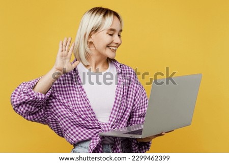 Young smiling happy fun blonde IT woman 20s she wears pink tied shirt white t-shirt hold use work on laptop pc computer get video call waving hand isolated on plain yellow background studio portrait Royalty-Free Stock Photo #2299462399