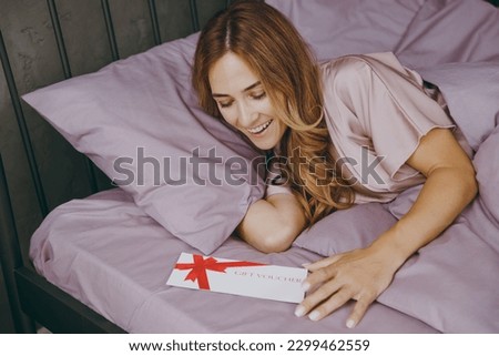 Young woman wear t-shirt pajama lying in bed hold gift certificate coupon voucher card for store rest relax spend time in bedroom lounge home in own room hotel wake up dream be lost in good mood day