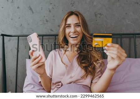 Young fun woman wear t-shirt pajama lying in bed using mobile cell phone credit bank card shopping online rest relax spend time in bedroom lounge home in hotel room wake up dream be lost in good mood