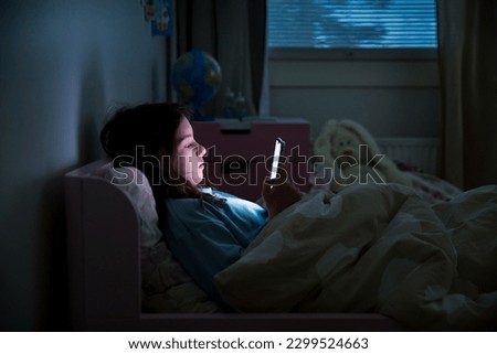 A child using smart phone lying in bed late at night, playing games, watching videos online, scrolling screen. Children's screen addiction. Child's room at night.  Royalty-Free Stock Photo #2299524663