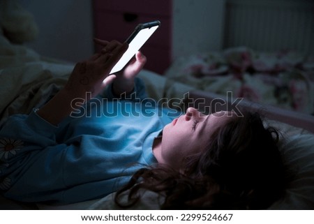 A child using smart phone lying in bed late at night, playing games, watching videos online, scrolling screen. Children's screen addiction. Child's room at night.  Royalty-Free Stock Photo #2299524667