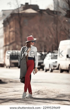 Stylish woman model in sunglasses walking in urban street wearing white shirt and red hat, looking away. Fashionable confident lady posing outdoors, lifestyle. Fashion style concept. Copy text space