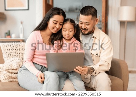 Asian Parents And Baby Daughter Using Laptop Watching Cartoons Online Discovering Digital World Together Sitting On Sofa At Home. Family Websurfing Via Computer Spending Quality Time On Weekend