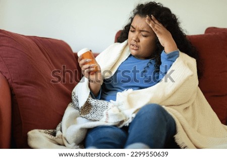 Sick sad young hispanic chubby curly woman in homewear sitting on couch covered in warm blanket, holding jar with drugs, touching her head, have headache, home interior, copy space Royalty-Free Stock Photo #2299550639