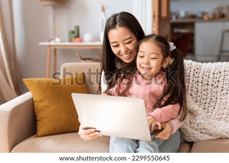 Cheerful Korean Mom And Baby Daughter Using Laptop Computer Engaging in Fun Online Activities, Watching Cartoons And Learning Together Sitting On Sofa At Home. Family Digital Leisure Concept