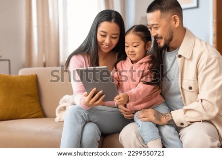 Family And Technology. Happy Asian Parents And Little Daughter Using Digital Tablet Computer, Surfing Internet And Watching Cartoons Online Via Gadget Sitting On Sofa At Home On Weekend