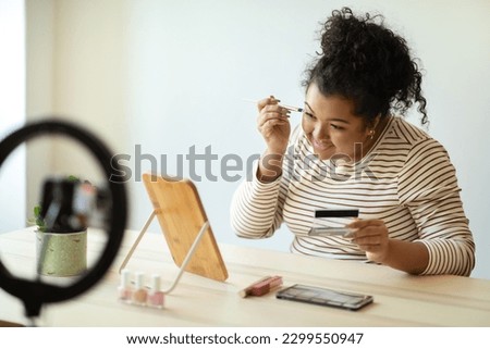 Beauty blog, blogging concept. Cheerful pretty curly young woman plus size in casual outfit sitting at desk in front of mirror at home, recording video while applying makeup, using blogger set