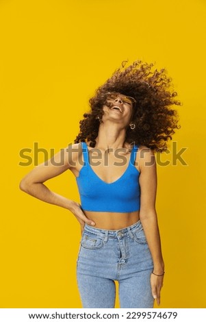 Woman with curly afro hair in a blue T-shirt on a yellow background dancing flying hair with sunglasses yellow, hand signs, look into the camera, smile with teeth and happiness, copy space