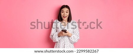 Online shopping. Amazed asian woman looking at camera with happy smile, making purchase with smartphone, using mobile phone app, standing over pink background.