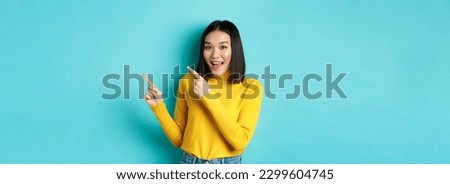 Shopping concept. Beautiful chinese girl in yellow sweater pointing fingers at upper right corner logo banner, smiling amused, standing over blue background.