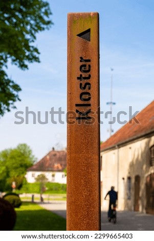 Sign outside Raitenhaslach monastery in Germany, indicating direction of entrance with a Maypole in the background.