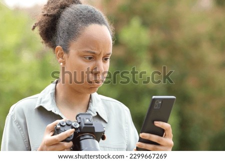 Frustrated black photographer holding mirrorless camera checking phone in a park