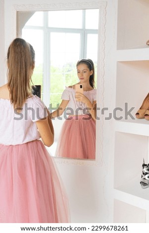 Rear view of tender teen girl in pink tutu-skirt and black high-heeled shoes with cell phone posing in front of mirror in wardrobe room, looking at own reflection and making selfie on smartphone