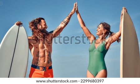 Caucasian woman in a swimsuit and a bearded muscular man with tattoos in swimming trunks with surfboards near the sea giving a high five gesture. Sports and active recreation. Royalty-Free Stock Photo #2299680397