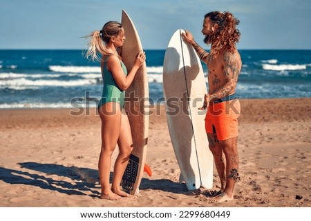 A couple of surfers on the beach. Caucasian woman in a swimsuit and a bearded buff man with tattoos in swimming trunks with surfboards near the sea. Sports and active recreation.