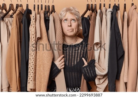 Horizontal shot of thoughtful blonde woman wears black jumper concentrated overhead poses among clohtes on hangers thinks what to wear chooses outfit from wardrobe. Women and fashion concept