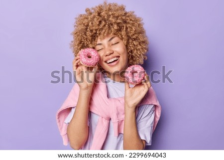 Tasty food and diet. Happy overjoyed curly haired woman holds delicious glazed doughnuts keeps eyes closed from pleasure dressed in t shirt and jumper tied over shoulders isolated on purple background Royalty-Free Stock Photo #2299694043