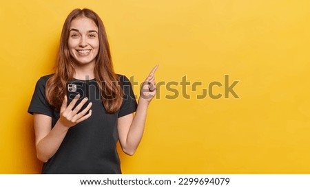 Horizontal shot of optimistic young European woman dressed in casual black t shirt holds mobile phone downloads new app points index finger above smiles toothily isolated over yellow background