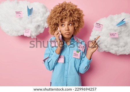 Busy sad curly haired woman makes phone call and points index finger aside wears shirt covered with sticky notes tries to decide on something stands indoor against pink background white clouds above