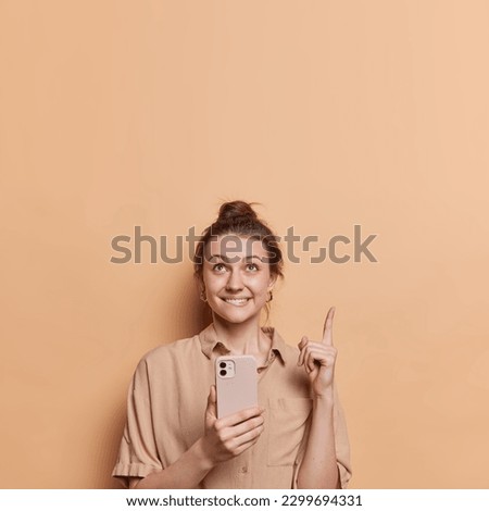 Click on this link. Positive woman with darkhair gathered in bun holds smartphone points index finger overhead demonstrates copy space wears shirt isolated over brown background. Technology concept Royalty-Free Stock Photo #2299694331