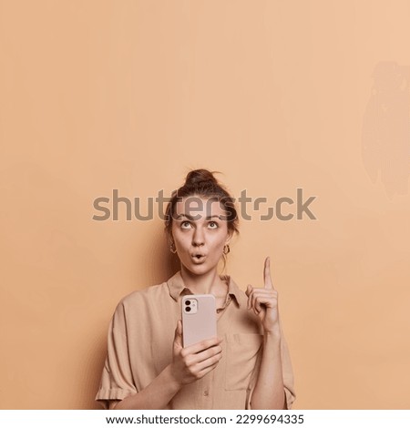 Stunned dark haired young woman looks with omg expression above points index finger on copy space holds smartphone asks to check out this promo dressed in shirt isolated over brown background.