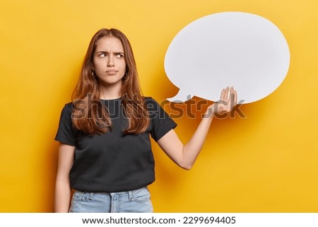 Horizontal shot of displeased long haired young woman frowns face holds speech bubble for your text being dissatified with situation dressed in casual clothes isolated over yellow background