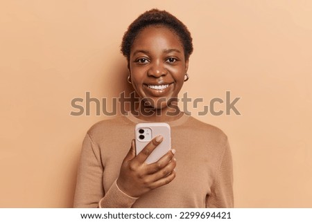 Portrait of dark skinned young pretty woman with short hair uses mobile device for communication browses web dressed in casual jumper smiles broadly has white teeth isolated over brown background.