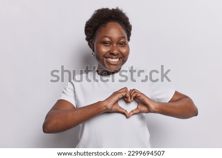 Lovely dark skined woman expresses sympathy and admiration makes heart gesture smiles toothily has romantic mood dressed in t shirt isolated over white background. Be my valentine. I love you sign