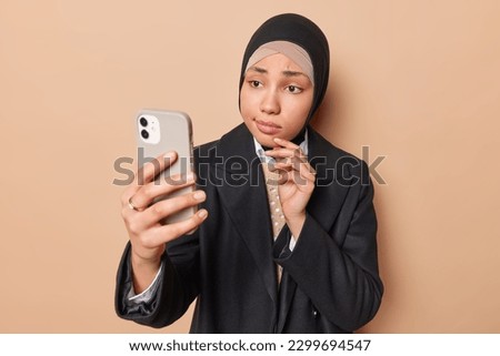 Photo of serious young Muslim woman takes selfie via smartphone dressed in formal clothes makes video call isolated over brown background. Good looking religious female model with modern gadget