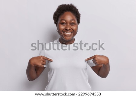 Place your advertising text here. Cheerful dark skinned plump woman points at blank space of t shirt for your logo or template smiles happily isolated over white background expresses positive emotions Royalty-Free Stock Photo #2299694553