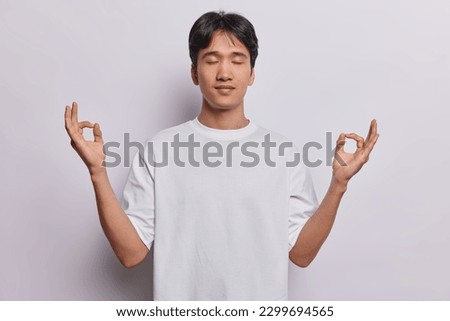 Handsome Chinese man with peaceful expression meditates indoor tries to calm down and relax on break practices yoga dressed in casual t shirt isolated on white background enjoys silence finds balance