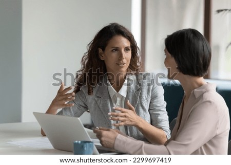 Two business women sit at desk discuss project details, diverse female colleagues met in office, share opinion, working on collaborative task, sales manager makes commercial offer to company client Royalty-Free Stock Photo #2299697419