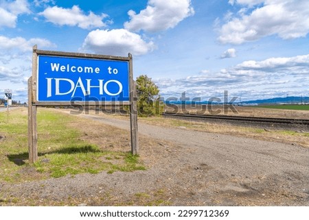 A roadside welcome to Idaho sign alongside a rural railroad track, coming from Spokane Washington and entering North Idaho, with the cities of Post Falls and Coeur d'Alene in view behind.