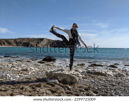 Full body photo of a white woman doing a yoga pose by the sea. Hand to big toe pose at Lulworth Cove, Dorset.