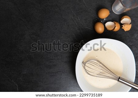 Pancake mix with egg, flour and milk with whisk on top and copy space on black stone countertop.