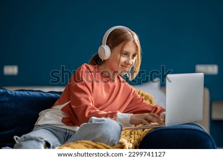 Smiling positive teen girl in headphones watching video movie on laptop sitting on couch at home. Leisure, recreation, entertainment pastime of teenager choosing film, cartoon to watch on computer.  Royalty-Free Stock Photo #2299741127