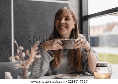 Portrait of joyful young woman enjoying a cup of tea and aroma at restaurant. Smiling pretty girl drinking hot tea in cafe. Excited blonde woman with bang wearing grey sweater and laughing. Royalty-Free Stock Photo #2299747517