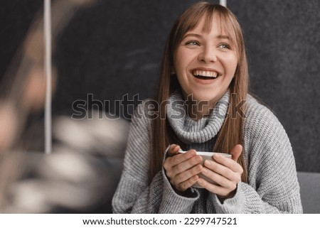 Portrait of joyful young woman enjoying a cup of tea and aroma at restaurant. Smiling pretty girl drinking hot tea in cafe. Excited blonde woman with bang wearing grey sweater and laughing.