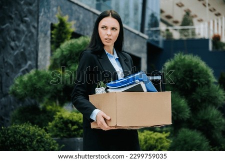 An office worker standing outside with a box, portraying the loss of their job and work. The Hardship of Joblessness Woman Holding Box After Dismissal Royalty-Free Stock Photo #2299759035
